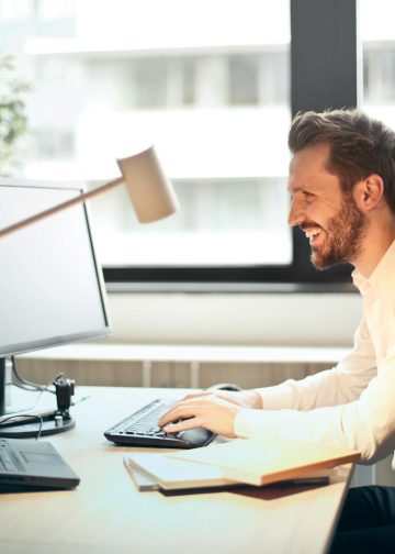 Man in White Dress Shirt Sitting on Black Rolling Chair While Facing Black Computer Set and Smiling